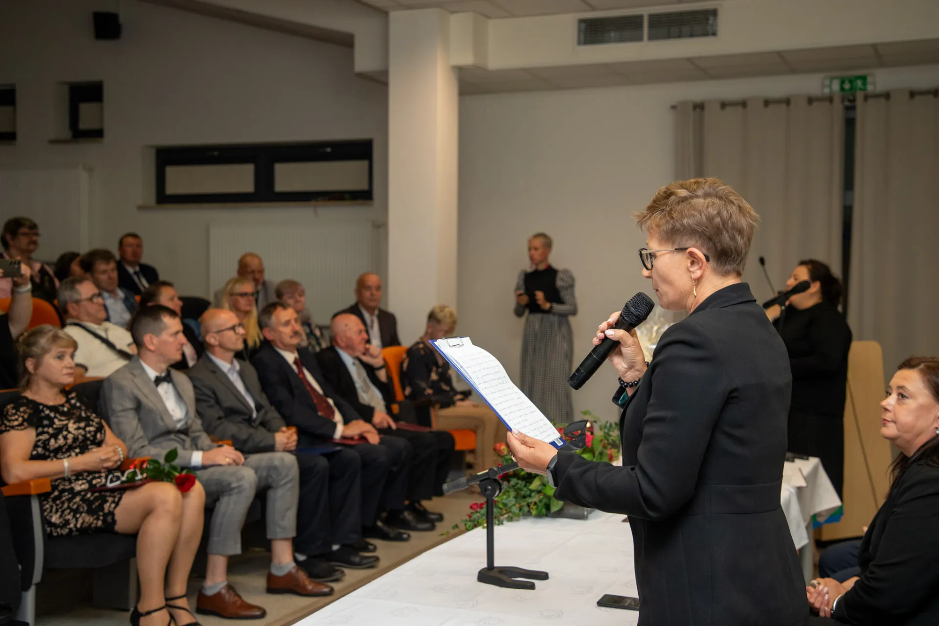 Na pierwszym planie stoi kobieta i czyta przemóienie, trzymając w ręku mikrofon, przed nią siedzą na widowni zaproszeni goście, uczestnicy Uroczystości