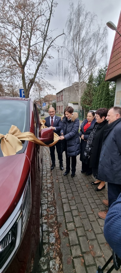 Pokaż zdjęcie: Trzy kobiety oraz dwóch mężczyzn stoi obok busa z dużą kokardą na jego przedniej szybie. Jedna z kobiet przecina wstęgę w kolorze złotym. W tle drzewa pozbawione liści oraz budynki mieszkalne.