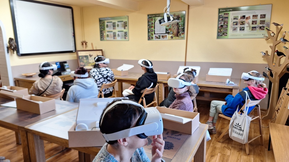 Warsztaty z udziałem okularów Virtual Reality. Grupa osób siedząca w sali edukacyjnej ma na głowie okulary Virtual Reality. W sali na ścianach tablica interaktywna, plansze edukacyjne ze zwierzętami.