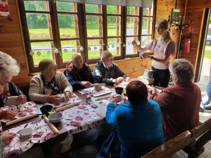 Warsztaty ceramiczne. Grupa starszych osób wykonuje prace ceramiczne siedząc przy stole w budynku. Instruktorka po lewej pokazuje gotowe prace.