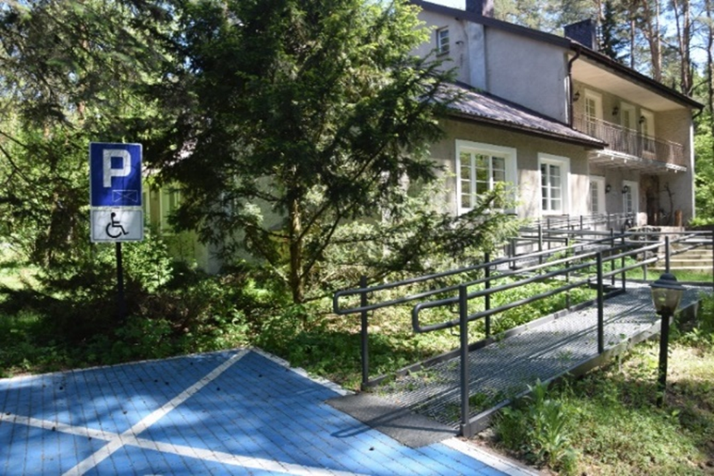 Miejsce parkindowe wraz z podjazdem do siedziby Mazowieckiego Parku Krajobrazowego. Miejsce parkingowe dla osób z niepełnosprawnościami, z którego do budynku w tle prowadzi metalowy podjazd. Wokół drzewa.