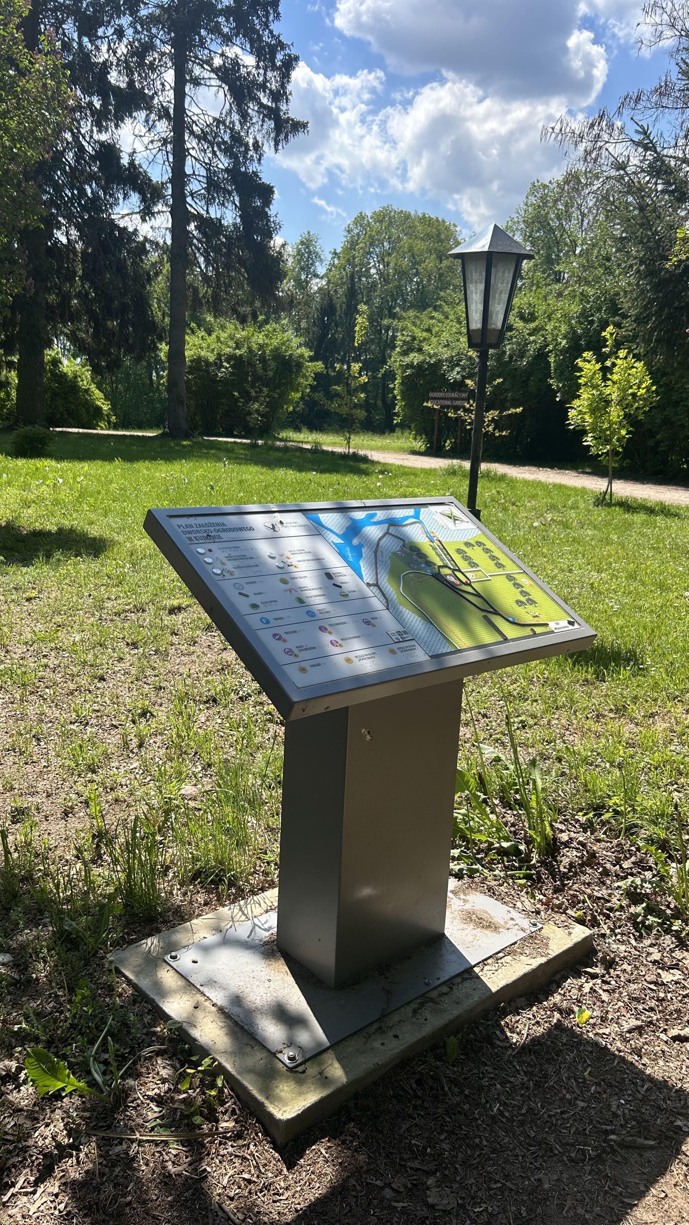 Plan tyflograficzny parku przydworskiego w Narwiańskim PN na metalowym postumencie. W tle latarnia, ścieżka leśna, trawa, drzewa i błękitne niebo.