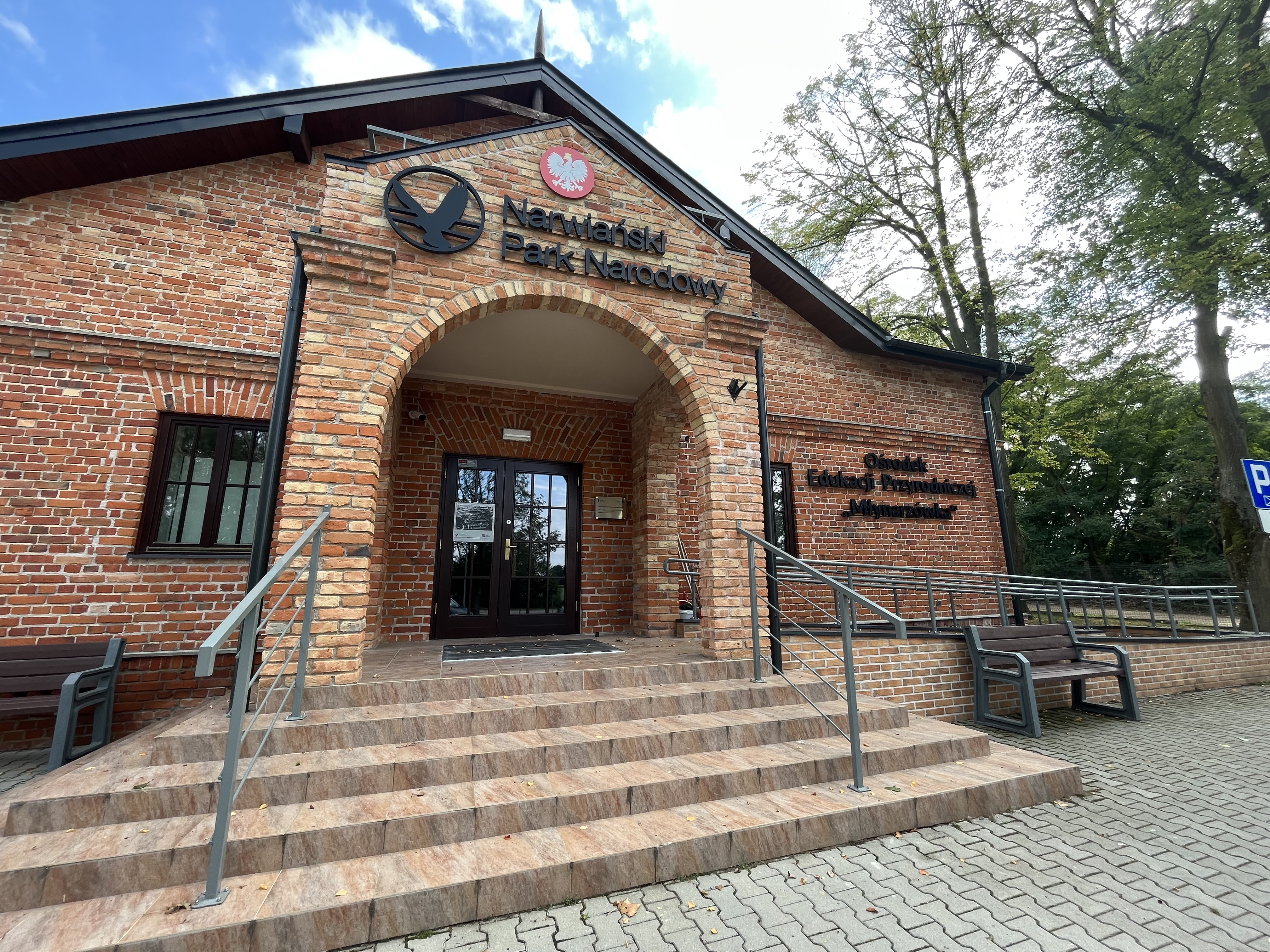 Na wprost schody z płytek i fasada budynku Ośrodka Edukacji Przyrodniczej "Młynarzówka" Narwiańskiego PN zrobiona z czerwonej cegły. Z prawej strony podjazd na wózków. Po obu stronach schodów ławki. W tle drzewa.