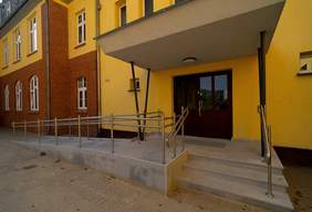 Pokaż zdjęcie: Podjazd wyłożony płytkami i otoczony metalową balustradą prowadzi do trzypiętrowego budynku. Do szkoły prowadzą także schody, a wejście jest zadaszone.