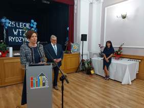 Pokaż zdjęcie: Po lewej stronie sali stoi kobieta przy mikrofonie. Z tyłu znajduje się mężczyzna w garniturze. W tle widoczny jest napis: „25-lecie WTZ w Rydzynie”. Po prawej stronie możemy dostrzec drugą kobietę opierającą się o stół.
