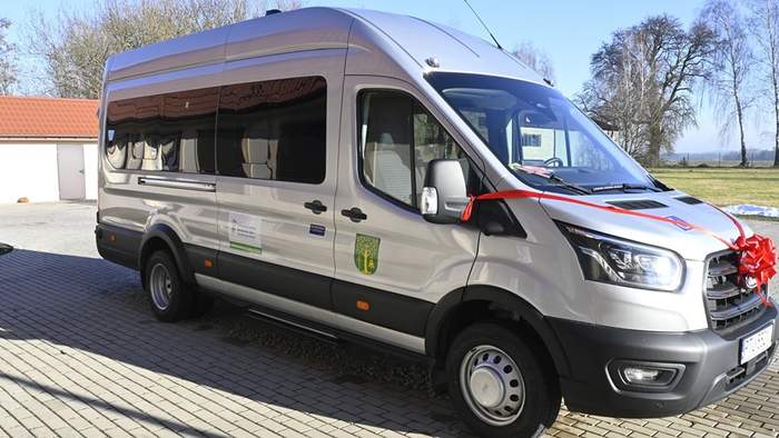 Pokaż zdjęcie: Srebrny autobus z czerwoną kokardą na masce pojazdu stoi zaparkowany na placu wyłożonym kostką brukową. W tle niski budynek z czerwoną dachówką, drzewa pozbawione liści oraz bezchmurne niebo.