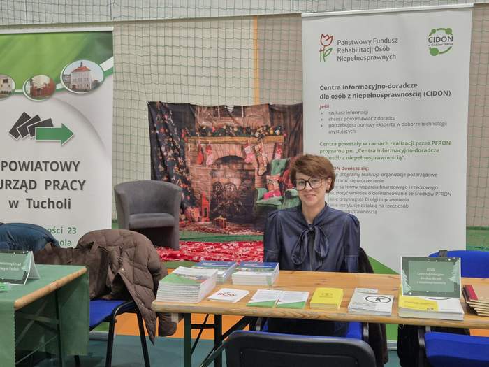 Punkt informacyjny CIDON PFRON - przy stoliki siedzi uśmiechnięta kobieta w okularach. Przed nią leżą ulotki i stoi koszyk z cukierkami. Za nią widać baner instytucji.
