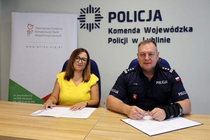 Pokaż zdjęcie: Przedstawiciel PFRON i funkcjonariusz Policji podpisują dokumenty przy stole w siedzibie Komendy Wojewódzkiej Policji w Lublinie, na tle logotypów instytucji.