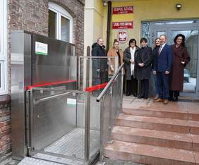 Pokaż zdjęcie: Zdjęcie przedstawia wejście do budynku szkoły z nowo zamontowanym podnośnikiem pionowym obok schodów. Po lewej stronie u dołu znajduje się pionowy podnośnik z przeszklonymi bokami i czerwonymi poziomymi listwami zabezpieczającymi.   Po prawej stronie są schody koloru czerwono-brązowego prowadzących do drzwi wejściowych. Przed wejściem do budynku, stoi siedem dorosłych osób w eleganckich ubraniach, ustawionych w lekkim półokręgu, patrzących w stronę aparatu.   Nad nimi, na żółtej ścianie budynku, wiszą czerwone tablice z białym napisem „Zespół Szkół w Obrzycku” oraz „Szkoła Podstawowa w Obrzycku”.