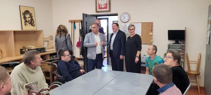 Pokaż zdjęcie: W jasnym pomieszczeniu, przy podwójnym stole siedzi kilka osób, w tym jedna na wózku inwalidzkim, przed stołem stoi trzech mężczyzn w marynarkach i dwie kobiety. Nad wejściem do pomieszczenia godło Polski po lewej wizerunek Chrystusa.