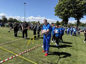 Pokaż zdjęcie: Uczestnicy zawodów sportowych na boisku biorą udział w przeciąganiu liny. Na pierwszym planie mężczyzna w niebieskim stroju sportowym i czerwonych butach trzyma linę. W tle zawodnicy, kibice i sędziowie pod słonecznym niebem.