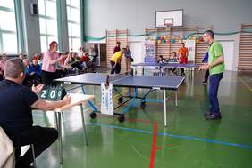 Pokaż zdjęcie: W hali sportowej trwa mecz tenisa stołowego. Po jednej stronie stołu kobieta w różowej bluzce przygotowuje się do serwisu, po drugiej mężczyzna w zielonej koszulce czeka na odbiór. W tle sędziowie i zawodnicy przy drugim stole, na trybunach siedzi publiczność. Na ścianie kolorowe balony i plakaty turnieju.