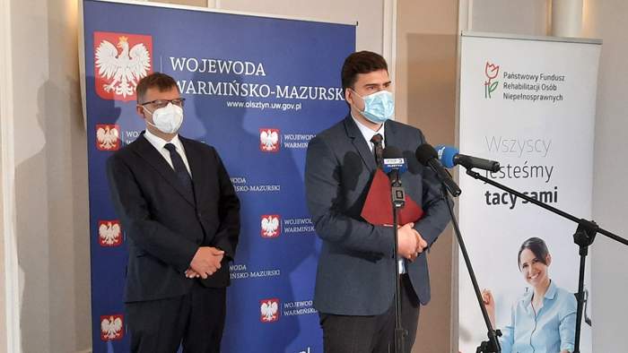 Pokaż zdjęcie: Dwóch panów w garniturach i maseczkach, przemawiających do niewidocznej na zdjęciu publiczności. Na tle banerów Wojewody i PFRON.  