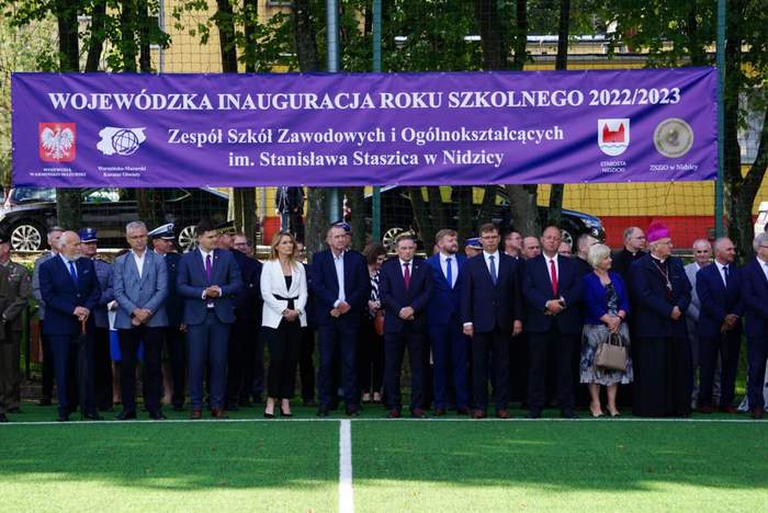 Pokaż zdjęcie: Zebrani przedstawiciele władz pod banerem z informacją o Wojewódzkiej Inauguracji Roku Szkolnego 2022/2023 z logami Województwa Warmińsko-Mazurskiego, Wojewódzkiego Kuratorium Oświaty, Starostwa Nidzickiego, ZSZiO w Nidzicy. 