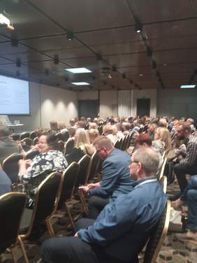 Pokaż zdjęcie: Uczestnicy konferencji podczas jednej z prezentacji, siedzą na krzesełkach ustawionych w długich rzędach.