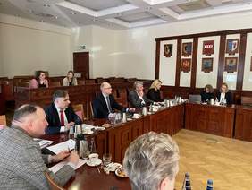 Pokaż zdjęcie: Za drewnianym rzeźbionym stołem w kształcie litery U siedzą na galowo ubrani ludzie. N a ścianach wiszą godła gmin pomorskich.