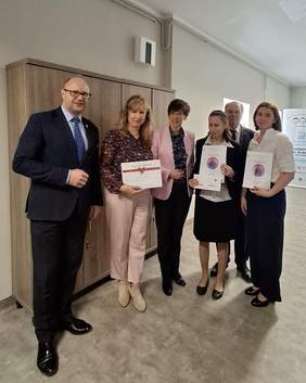 Pokaż zdjęcie: Grupowe zdjęcie sześciu osób stojących w holu Warsztatu Terapii Zajęciowej w Gołańczy. Trzy osoby trzymają w dłoniach dyplomy oraz bon podarunkowy. Goście ubrani są elegancko w garnitury, marynarki oraz eleganckie bluzki. Uroczyste zdjęcie z okazji wygranej pani Grażyny Wleczyk laureatki III miejsca w ogólnopolskim Konkursie Plastycznym „Sztuka Osób Niepełnosprawnych”.