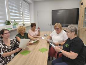 Pokaż zdjęcie: Pięć kobiet siedzi przy stole w sali konferencyjnej, czytając dokumenty podczas spotkania roboczego w Ośrodku Wsparcia i Testów w Białymstoku.