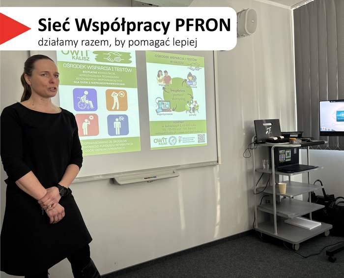 Pokaż zdjęcie: W centralnej części slajdu widać kobietę w czarnym stroju, stojącą przy tablicy projektowej i mówiącą do grupy. W tle jest wyświetlana prezentacja na tablicy. Po prawej stronie sali stoi wózek ze sprzętem i monitor, a przy oknie są zasłony. Na slajdzie w lewym górnym rogu jest biały pasek z czerwonym trójkątem i dużym napisem: „Sieć Współpracy PFRON”, a pod nim mniejszy: „działamy razem, by pomagać lepiej”.