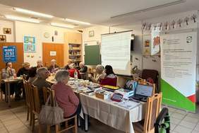 Pokaż zdjęcie: Spotkanie z seniorami w Krakowskim Centrum Seniora – Dom nr 7. Grupa seniorów siedzi przy stole. Obok stołu stoją przedstawiciele PFRON i OWiT, opowiadają o formach wsparcia. Przy stole postawione są rollupy z logo PFRON i OWiT.