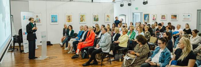 Pokaż zdjęcie: Sala konferencyjna z drewnianą podłogą i białymi ścianami z zawieszonymi ilustracjami dzieci. Kobieta w czarnym garniturze przemawia przy mównicy z mikrofonem do siedzącej publiczności. 