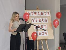Pokaż zdjęcie: Kobieta przemawia przy mównicy podczas gali „Otwarte Serca Roku 2026”. W tle tablica z nazwą wydarzenia, czerwone balony i statuetki przygotowane dla wyróżnionych. Sala udekorowana w biało-czerwonych barwach.