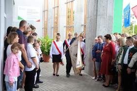 Pokaż zdjęcie: Sztandar szkoły niosło troje uczniów w galowych strojach. Ceremonii przyglądali się uczniowie i zaproszeni goście. Na budynku widoczna jest tablica z logiem i dofinansowaniem PFRON.  