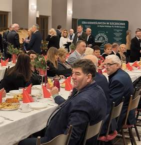 Pokaż zdjęcie: Osoby siedzące przy stole z białymi obrusami, na którym znajdują się czerwone serwetki i poczęstunek, w tle osoby stojące przy zielonym banerze z białymi napisami "Izba Rzemieślnicza w Szczecinie"