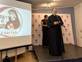 Pokaż zdjęcie: Duchowny w czarnej sutannie przemawia do mikrofonu na scenie. Za nim stoi kobieta w czarnej sukni. Obok wyświetlana jest grafika Centrum Pomocowego Caritas z wizerunkiem św. Siostry Faustyny.