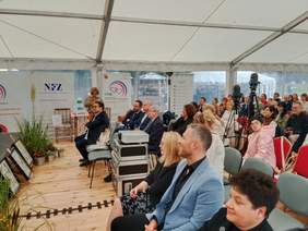 Pokaż zdjęcie: Na krzesłach siedzą ludzie. Obserwują to, co dzieje się na scenie. W tle widać także ustawione kamery. 
