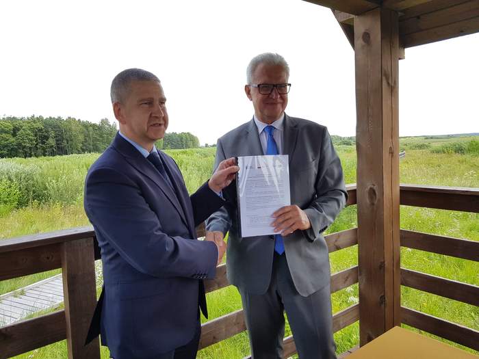 Pokaż zdjęcie: 2 mężczyzn w garniturach uściskujących sobie dłonie w geście gratulacji. Wspólnie prezentują trzymany przez siebie dokument. W tle zielony plener.