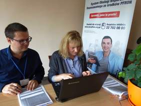 Pokaż zdjęcie: Dwie osoby siedzą przy stoliku, na którym leży laptop i dokumenty