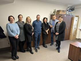 Pokaż zdjęcie: Biuro. Na zdjęciu dwóch mężczyzn i dwie kobiety, Wszyscy pozują do zdjęcia i się uśmiechają. Za nimi ciemne szafki. Stoją na szarej wykładzinie.