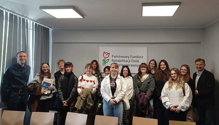 Pokaż zdjęcie: Grupa kilkunastu osób stoi w sali konferencyjnej na tle dużego logotypu PFRON zawieszonego na ścianie. Z przodu znajduje się rząd sześciu krzeseł. Okna przysłonięte są żaluzjami. Na suficie podwieszone są dwie lampy, które w danej chwili są włączone.
