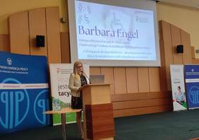 Pokaż zdjęcie: Kobieta w jasnej marynarce i czarnej sukience przemawia przy mównicy w sali konferencyjnej. Za nią wyświetlony jest slajd z prezentacją Barbary Engel, Zastępcy Prezesa Zarządu ds. Finansowych Państwowego Funduszu Rehabilitacji Osób Niepełnosprawnych, zatytułowaną: 'Od wsparcia do samodzielności – aktywizacja zawodowa jako klucz do niezależności osób z niepełnosprawnościami'. W tle stoją banery promujące PFRON oraz Państwową Inspekcję Pracy