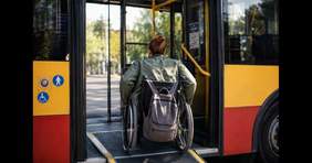 Pokaż zdjęcie: Do autobusu wjeżdża kobieta siedząca na wózku inwalidzkim. Na oparciu wózka inwalidzkiego przewieszony jest plecak. W tle widać wnętrze autobusu. 