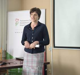 Pokaż zdjęcie: Kobieta z krótkimi włosami w ciemnej bluzce i jasnej spódnicy prowadzi wykład.