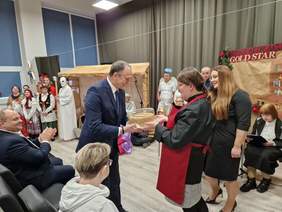 Mężczyzna otrzymuje od pani w czerwonym fartuszku tort, obok niej inna w czarnej sukience z długimi włosami, uśmiechają się, w tle uczestnicy przedstawienia w strojach jasełkowych i ludowych