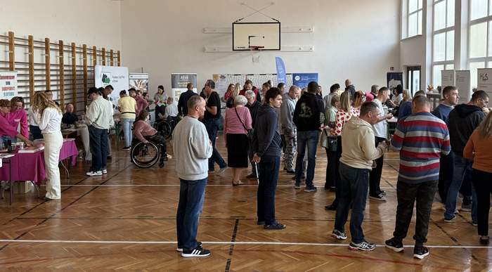 Pokaż zdjęcie: Spotkanie w sali gimnastycznej z drewnianą podłogą i wysokimi oknami, gdzie liczne osoby rozmawiają przy stoiskach informacyjnych z banerami i materiałami, obecne są także osoby na wózkach.