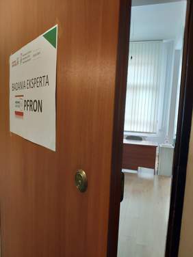 Pokaż zdjęcie: Na pierwszym planie są brązowe drzwi z naklejonym plakatem informującym o badaniach eksperta PFRON drzwi są uchylone widać kawałek jasnego biurka szafkę i duże okno zasłonięte białymi pionowymi żaluzjami