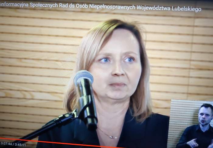 Pokaż zdjęcie: Przy mikrofonie stoi kobieta blondynka, zdjęcie popiersia, w prawym dolnym rogu widać tłumacza języka migowego