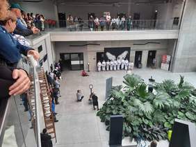 Pokaż zdjęcie: W holu dzieci i młodzież prezentują pokaz karate. Wokół nich z każdej strony stoją ludzie, którzy przyglądają się temu widowisku. 