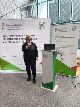 Pokaż zdjęcie: Kobieta z mikrofonem w ręku wita gości stojąc przy mównicy podczas wydarzenia PFRON. W tle stoją roll‑upy informacyjne.