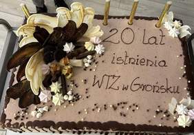 Pokaż zdjęcie: Tort w beżowym kolorze z napisem: „20 lat istnienia WTZ w Grońsku”. Z lewej strony tortu widoczne są dekoracje przestrzenne. Na torcie ustawione są świeczki zimne ognie, które nie zostały jeszcze zapalone.