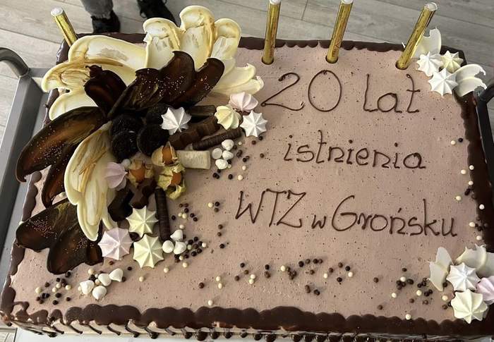 Pokaż zdjęcie: Tort w beżowym kolorze z napisem: „20 lat istnienia WTZ w Grońsku”. Z lewej strony tortu widoczne są dekoracje przestrzenne. Na torcie ustawione są świeczki zimne ognie, które nie zostały jeszcze zapalone.