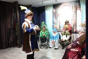 Pokaż zdjęcie: Grupa osób w kostiumach historycznych odgrywa scenę teatralną podczas jubileuszu 20-lecia Warsztatu Terapii Zajęciowej w Siemiatyczach. Na pierwszym planie mężczyzna w stroju królewskim z koroną w rękach, w tle inni uczestnicy w kolorowych strojach z perukami i nakryciami głowy.