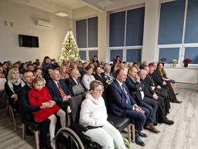 Zebrani goście, siedzą na krzesłach, za nimi w kącie widać ubraną choinkę