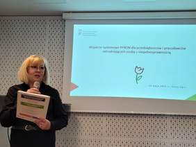 Pokaż zdjęcie: Dyrektor Marta Mordarska przemawia do mikrofonu. Za nią wyświetlane są slajdy prezentacji z logo PFRON.