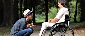 Pokaż zdjęcie: Po prawej stronie znajduje się kobieta, która siedzi na ręcznym wózku inwalidzkim. Po lewej stronie kuca przed nią mężczyzna. Oboje się uśmiechają do siebie.W tle widać drzewa.  