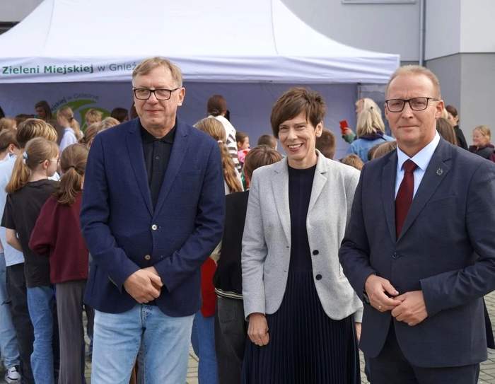 Pokaż zdjęcie: Oficjalne wydarzenie plenerowe. Od lewej stoją: Marek Machiński - dyrektor Szkoły Podstawowej nr 8 w granatowej marynarce, Anna Skupień - dyrektor Oddziału Wielkopolskiego PFRON w szarym żakiecie oraz prezydent Miasta Gniezna Michał Powałowski w garniturze z bordowym krawatem. W tle grupa młodzieży, biały namiot i ściana budynku.